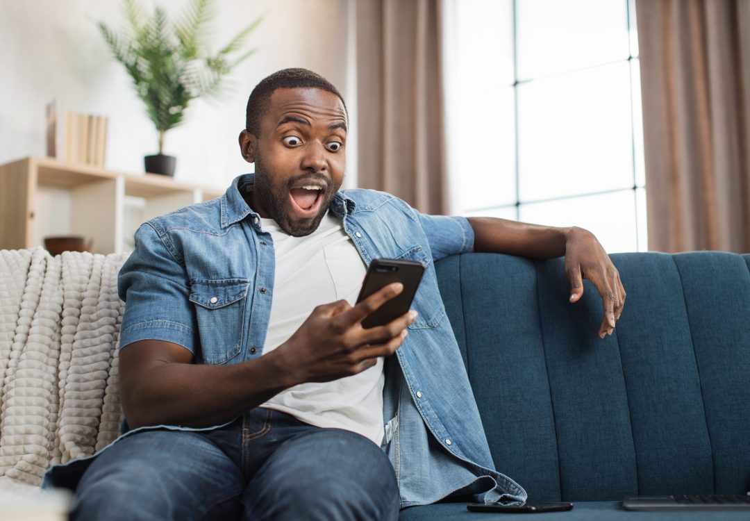 African american man in casual wear looking on mobile screen with amazed facial expression. Happy young guy sitting on couch amd reading good news on mobile.